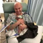 A smiling father holding a premature infant in a hospital room.