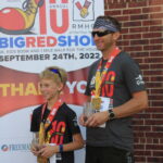 A father and son, wearing matching t-shorts and holding medals, stand next to each other after finishing a Big Red Shoe race.